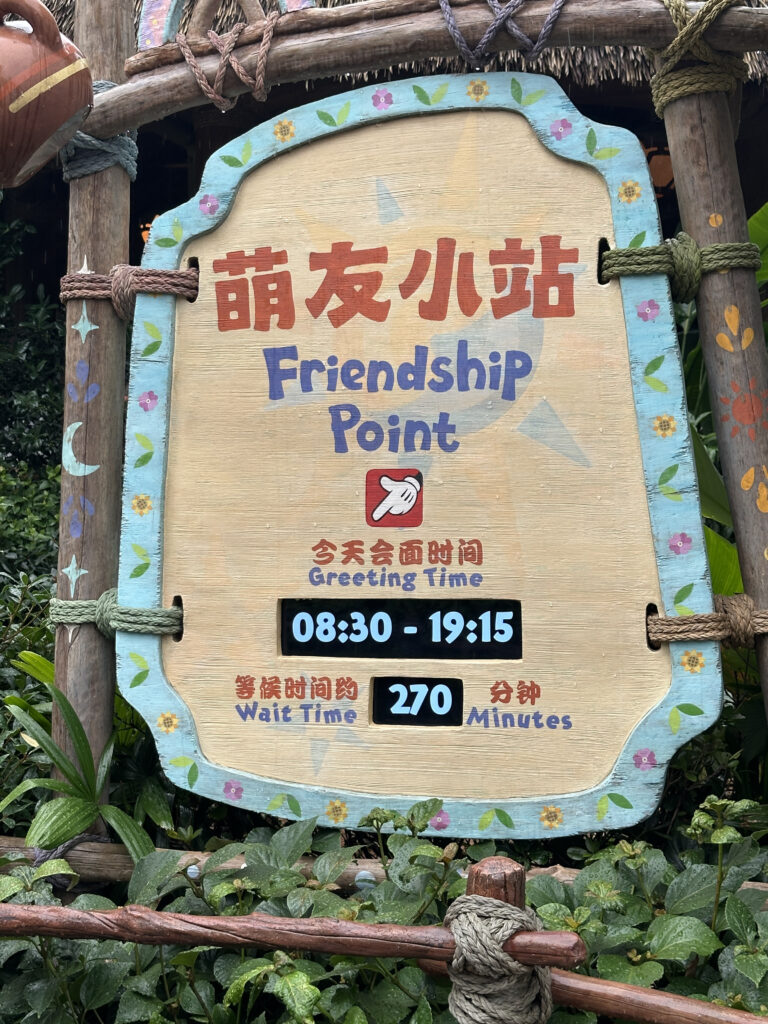 The image shows a wooden sign with decorative elements, including flowers and ropes. The sign reads "Friendship Point" in both Chinese and English. Below, it displays "Greeting Time" with the hours "08:30 - 19:15" and "Wait Time" as "270 Minutes." The sign is surrounded by greenery.