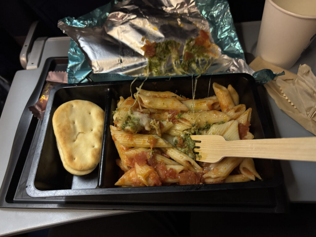 The image shows a meal served in a black plastic tray, likely on an airplane. The tray contains penne pasta with tomato sauce and herbs, and a piece of flatbread. A wooden fork is placed on the pasta. There is also a foil wrapper and a paper cup nearby.