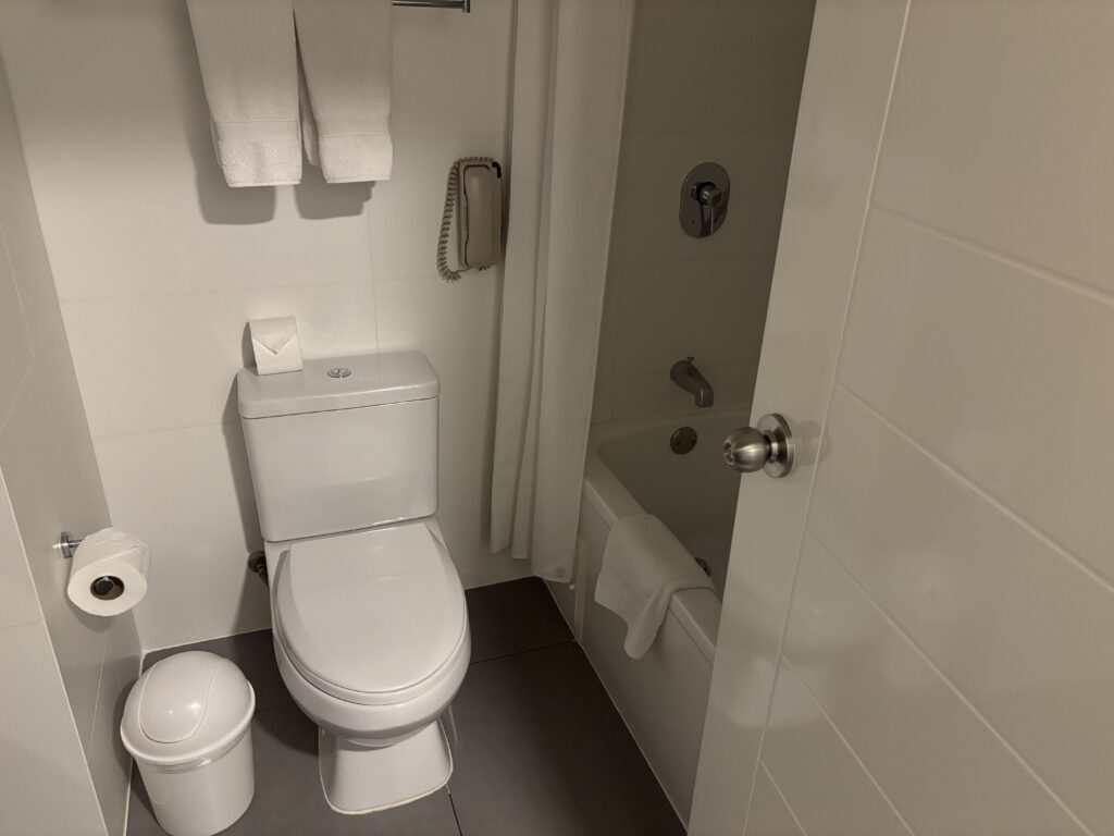 The image shows a clean, modern bathroom. There is a white toilet with a folded toilet paper on top of the tank. A roll of toilet paper is mounted on the wall, and a small trash bin is placed next to the toilet. Above the toilet, two white towels are hanging on a rack. To the right, there is a bathtub with a towel draped over the edge. A shower curtain is partially visible, and a wall-mounted telephone is next to the bathtub. The bathroom has white tiles and a gray floor.