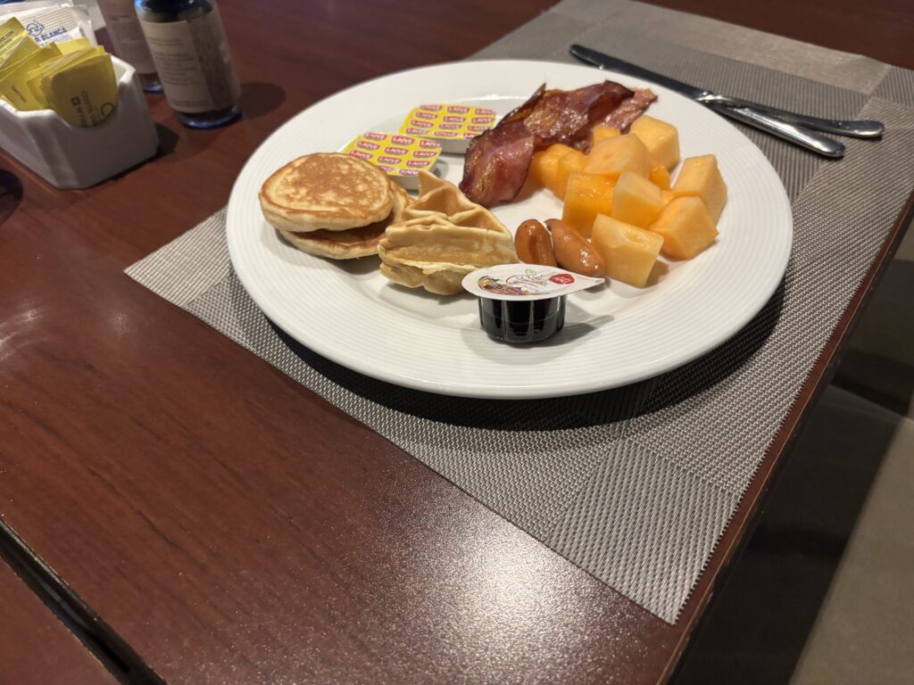 A breakfast plate on a table with a variety of foods, including small pancakes, bacon, cantaloupe cubes, sausages, and a container of syrup. There are also butter packets on the plate. The table is set with a placemat, and there are condiments and utensils nearby.