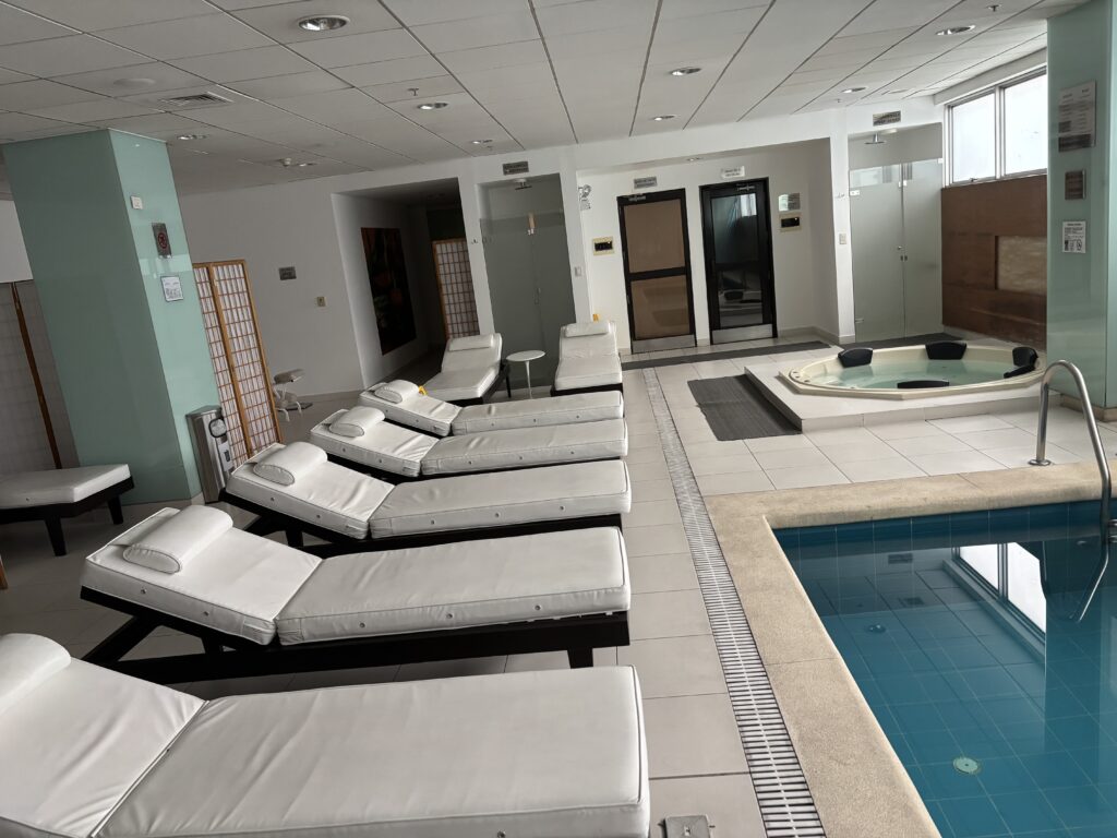 The image shows an indoor spa area with several white cushioned lounge chairs arranged in a row. There is a small pool in the foreground and a hot tub in the background. The room has a modern design with light-colored tiles and a ceiling with recessed lighting. There are also some folding screens and doors visible in the background.