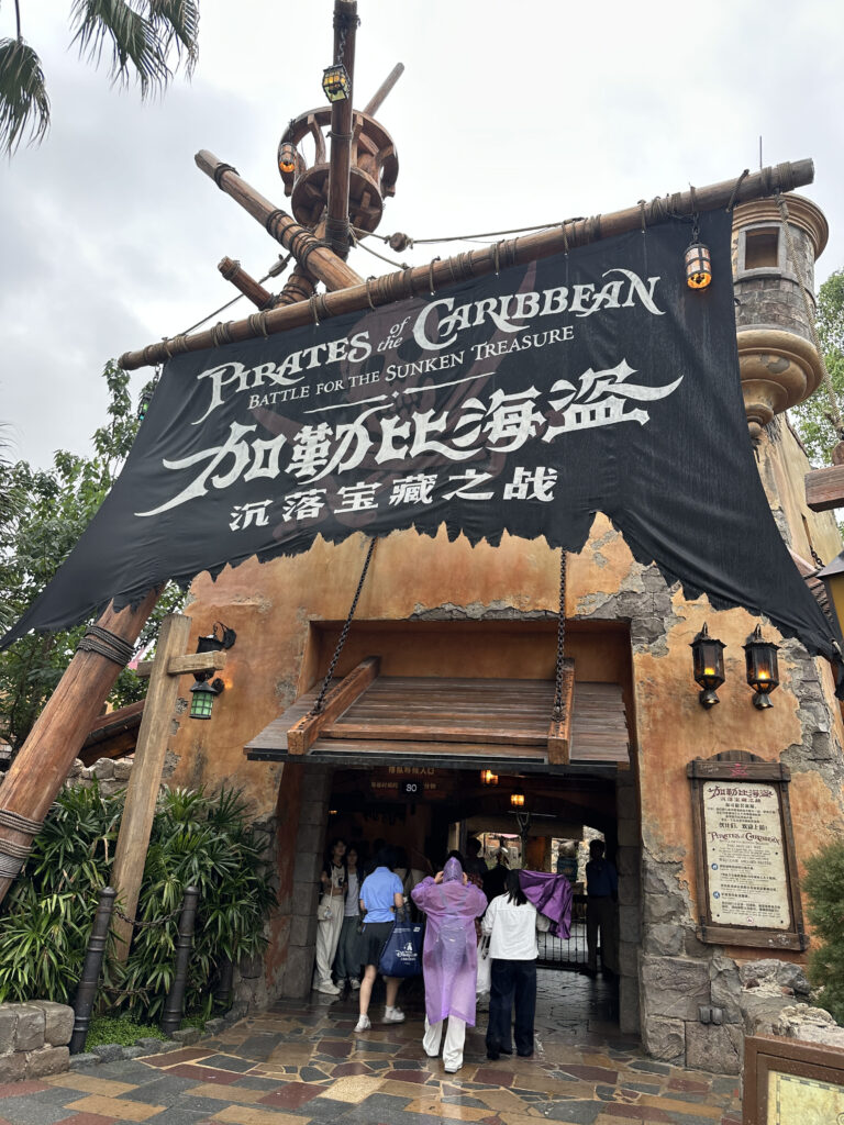The image shows the entrance to a theme park attraction called "Pirates of the Caribbean: Battle for the Sunken Treasure." The entrance is designed to look like a pirate ship with a large black sail featuring the attraction's name in both English and Chinese. Several people are entering the attraction, some wearing rain ponchos. The surrounding area is decorated with plants and stone walls, contributing to the pirate theme.