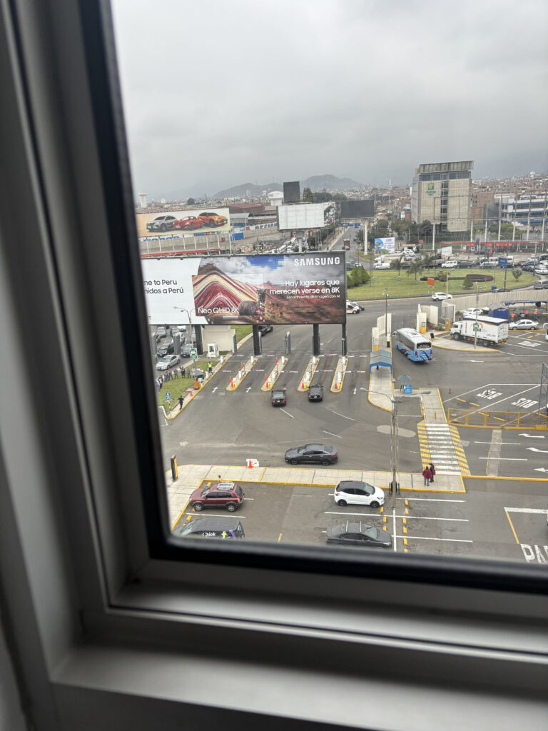 The image shows a view from a window overlooking a parking lot and a road. There are several cars parked and driving, with a few people walking. In the background, there are large billboards, one of which advertises Samsung Neo QLED 8K. The sky is overcast, and there are buildings and a cityscape visible in the distance.