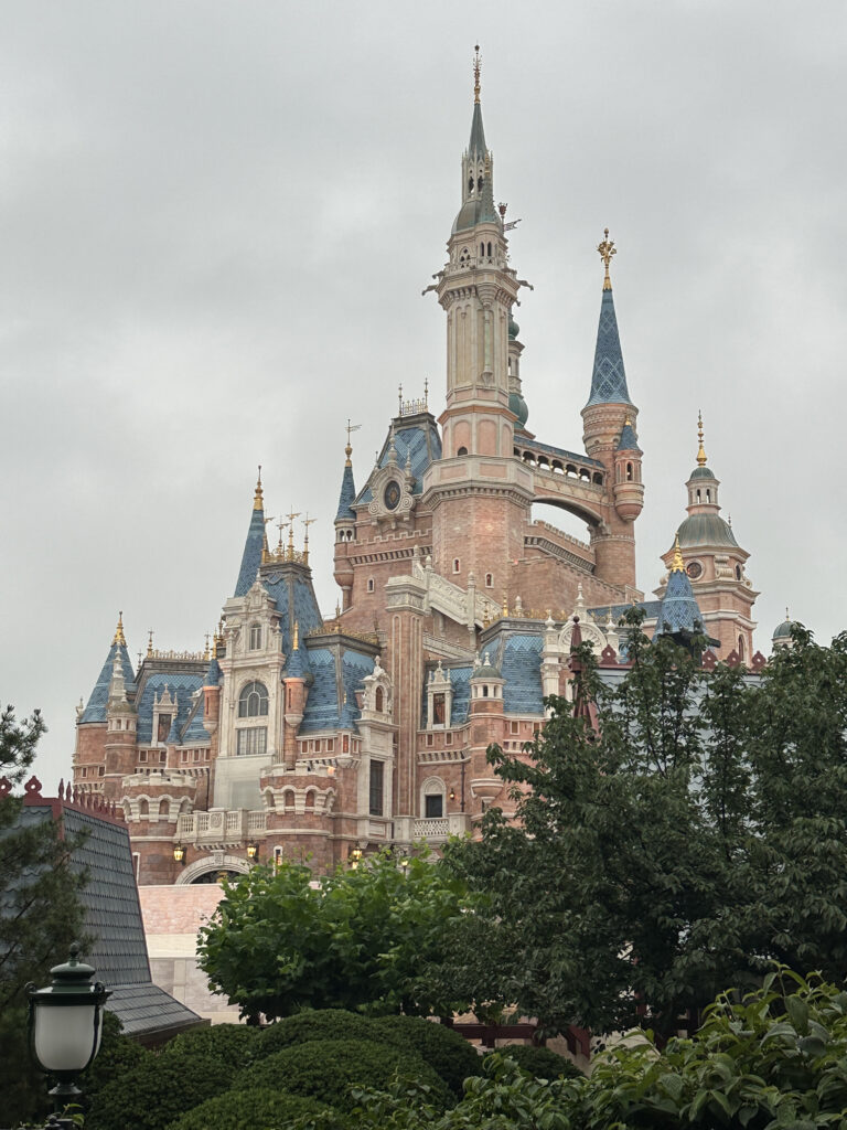 A large, ornate castle with multiple spires and turrets, featuring a mix of blue and gold accents. The castle is surrounded by lush green trees and bushes, and the sky is overcast.