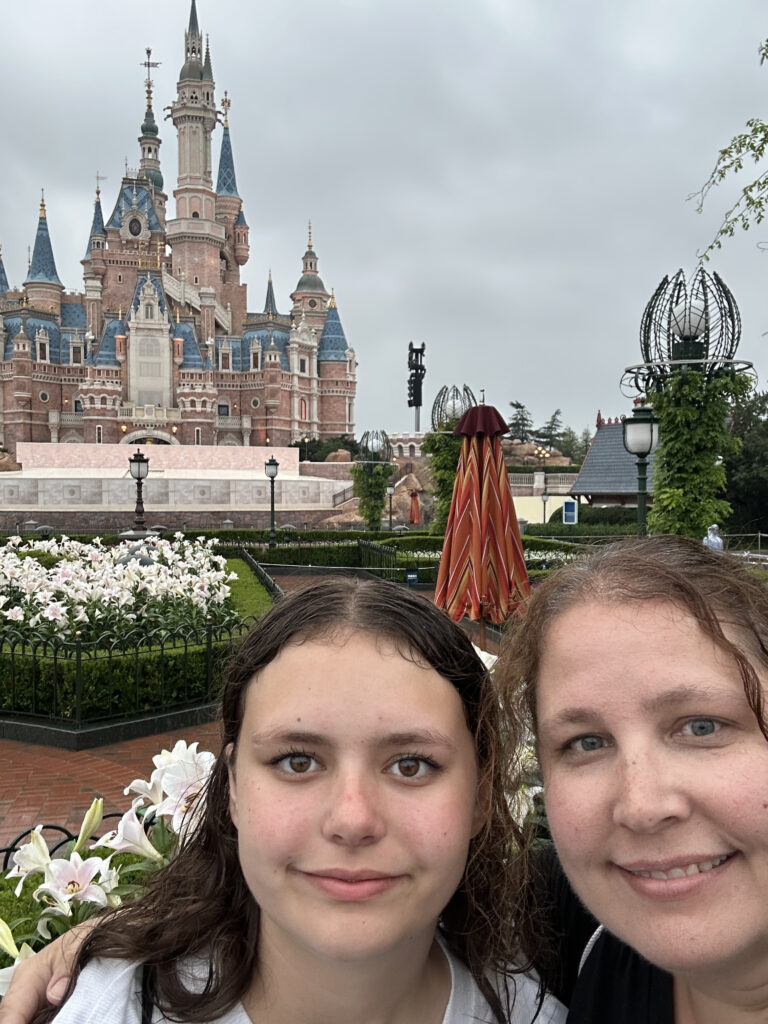 A selfie of two people in front of a large, ornate castle with blue and gold spires, resembling a Disney castle. The background includes a garden with white flowers and a cloudy sky.