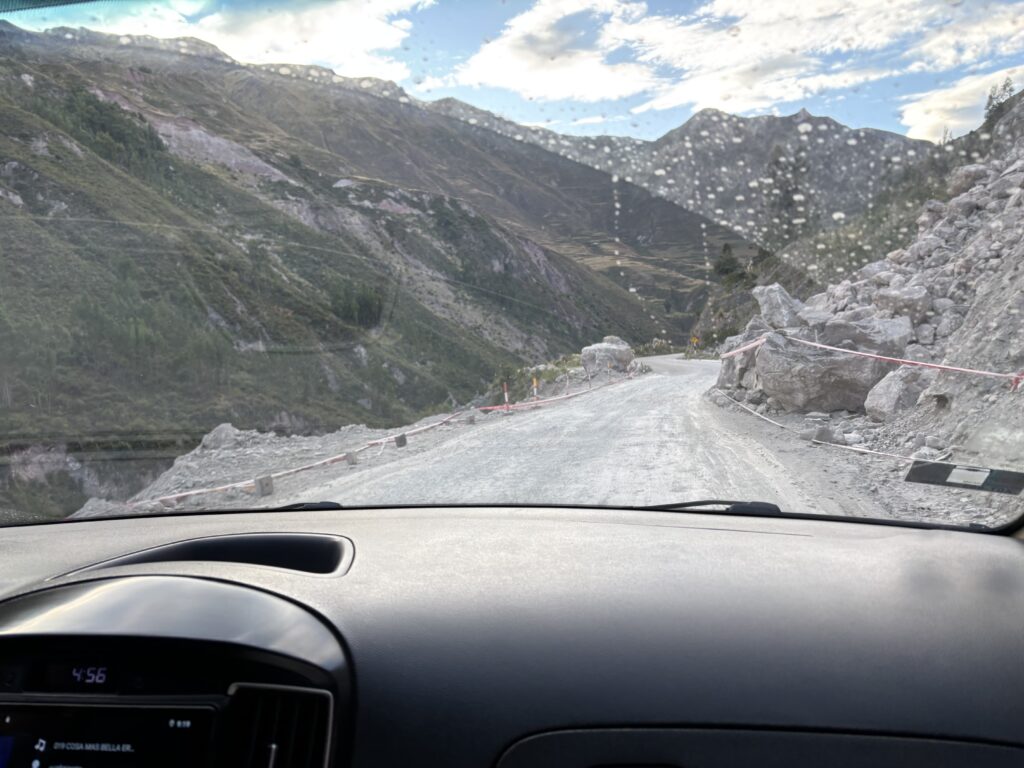 The image shows a view from inside a car driving on a narrow, winding mountain road. The road is unpaved and appears to be under construction, with large rocks and boulders on the side. There are red and white caution tapes along the edge. The windshield has water droplets on it, and the surrounding landscape features steep, green hills and mountains under a partly cloudy sky.