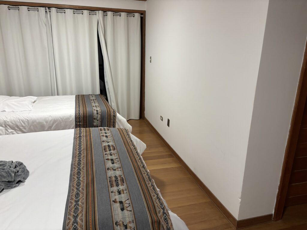 The image shows a room with two neatly made beds, each with a decorative runner featuring a geometric and animal pattern. The beds are positioned against a wall with white curtains, partially open, revealing a dark space behind them. The floor is wooden, and there is a plain white wall on the right side of the image. A small piece of gray fabric is on one of the beds.