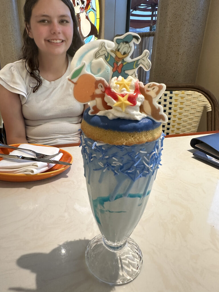 A girl is sitting at a table with a large, decorative milkshake in front of her. The milkshake is in a tall glass with blue sprinkles around the rim and blue swirls inside. It is topped with whipped cream, a donut, and colorful character-themed decorations, including figures resembling cartoon characters. There is a plate with a napkin and utensils on the table.