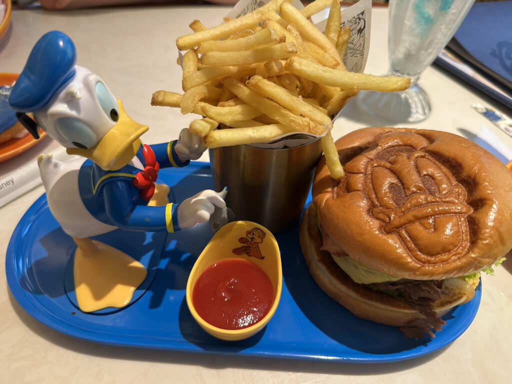 A blue tray holds a meal featuring a burger with a bun embossed with a cartoon character's face. Next to it is a metal cup filled with French fries. A small dish of ketchup is also on the tray. A figurine of a cartoon duck in a sailor outfit is positioned as if interacting with the fries. In the background, there is a glimpse of a milkshake or drink in a tall glass.