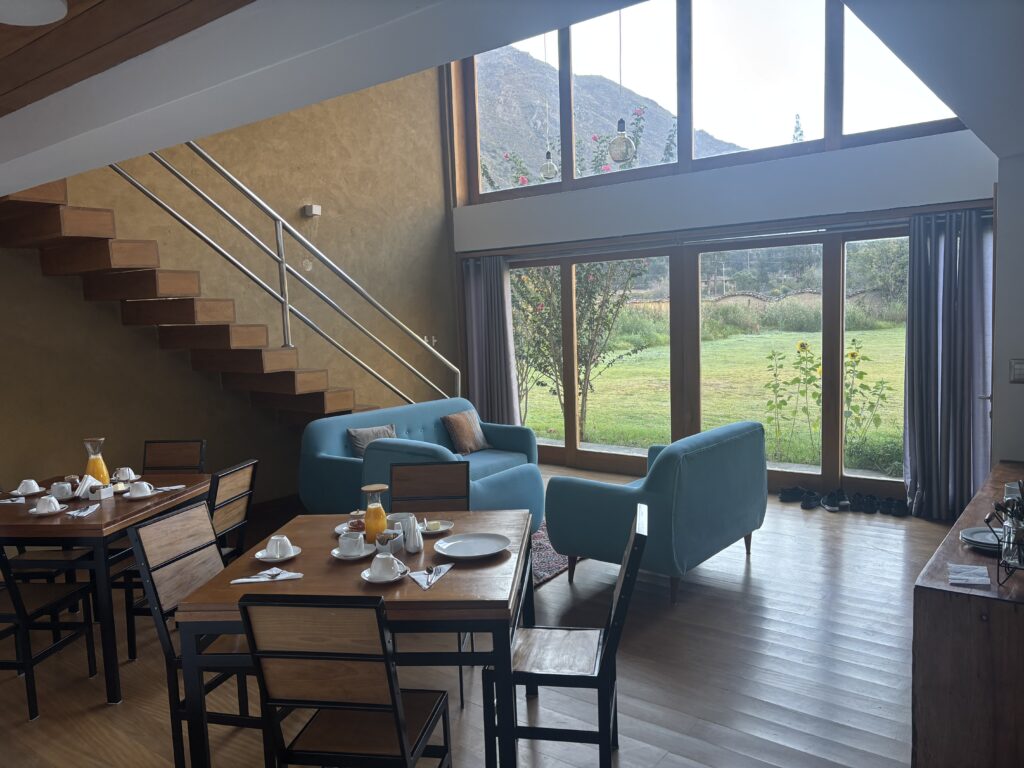The image shows a modern living and dining area with large windows offering a view of a grassy outdoor space and mountains in the background. Inside, there are two blue sofas and a wooden dining table set with white dishes and glasses of orange juice. A wooden staircase with a metal railing leads to an upper level. The room has wooden flooring and a warm, inviting atmosphere.