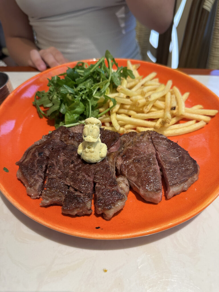 A plate of food featuring sliced steak with a dollop of butter on top, accompanied by a side of French fries and a small salad of leafy greens. The plate is orange, and a person is partially visible in the background.