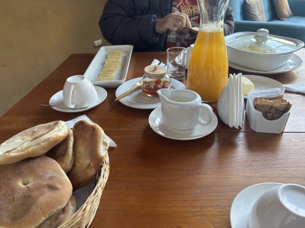 A breakfast table setting with various items. There is a basket of bread, a white teapot, a small pitcher of milk, and a cup and saucer. A glass jar of honey with a wooden dipper is present, along with a stack of butter slices on a rectangular plate. A carafe of orange juice is also on the table, along with a covered dish. Napkins and sugar packets are neatly arranged in holders. A person in a dark jacket is seated at the table.