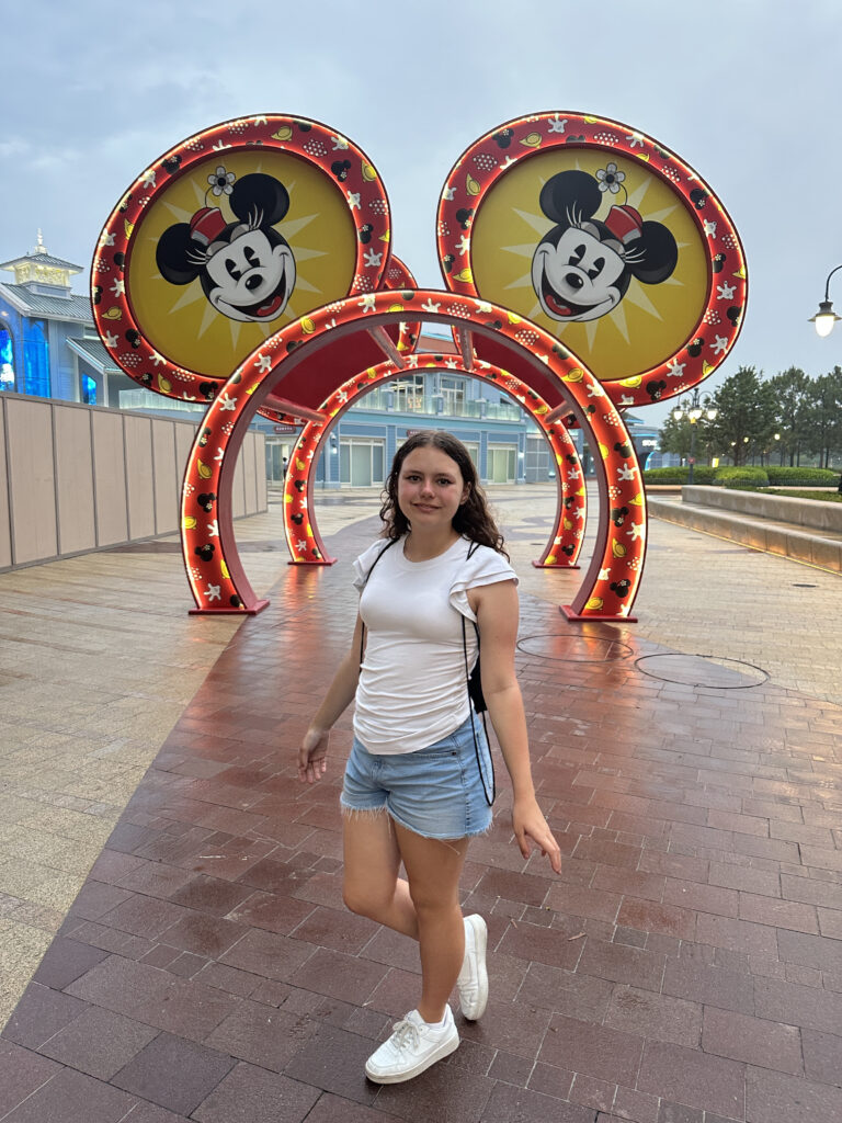 A young person is standing on a wet, tiled walkway, posing with one foot slightly raised. They are wearing a white t-shirt, denim shorts, and white sneakers. Behind them is a decorative archway featuring large circular designs with images of a cartoon mouse character. The archway is adorned with red and yellow colors and is lit with small lights. The background includes a building and some trees under a cloudy sky.