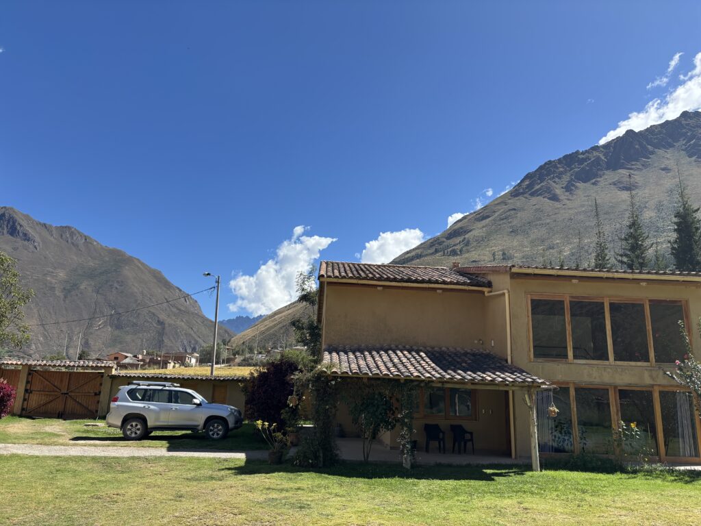 A two-story house with a tiled roof is situated in a grassy area. A silver SUV is parked nearby. In the background, there are large mountains under a clear blue sky with a few clouds.