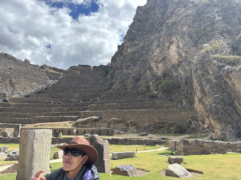 A person wearing a brown hat and sunglasses is in the foreground of the image, with a large stone structure and terraces in the background. The structure is built into a steep hillside, and there are people walking on the terraces. The sky is partly cloudy.