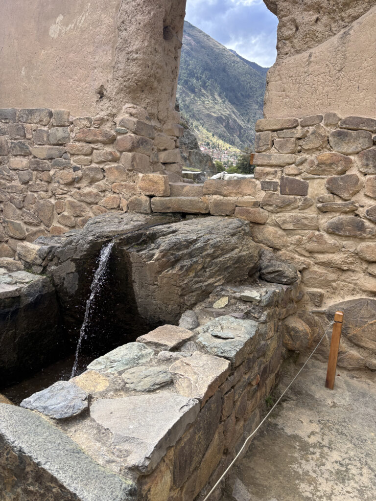 The image shows an ancient stone structure with a small stream of water flowing through a channel in the stones. The walls are made of rough, uneven stones, and there is an opening that reveals a mountainous landscape in the background. The sky is partly cloudy, and there is a rope barrier on the right side of the image.
