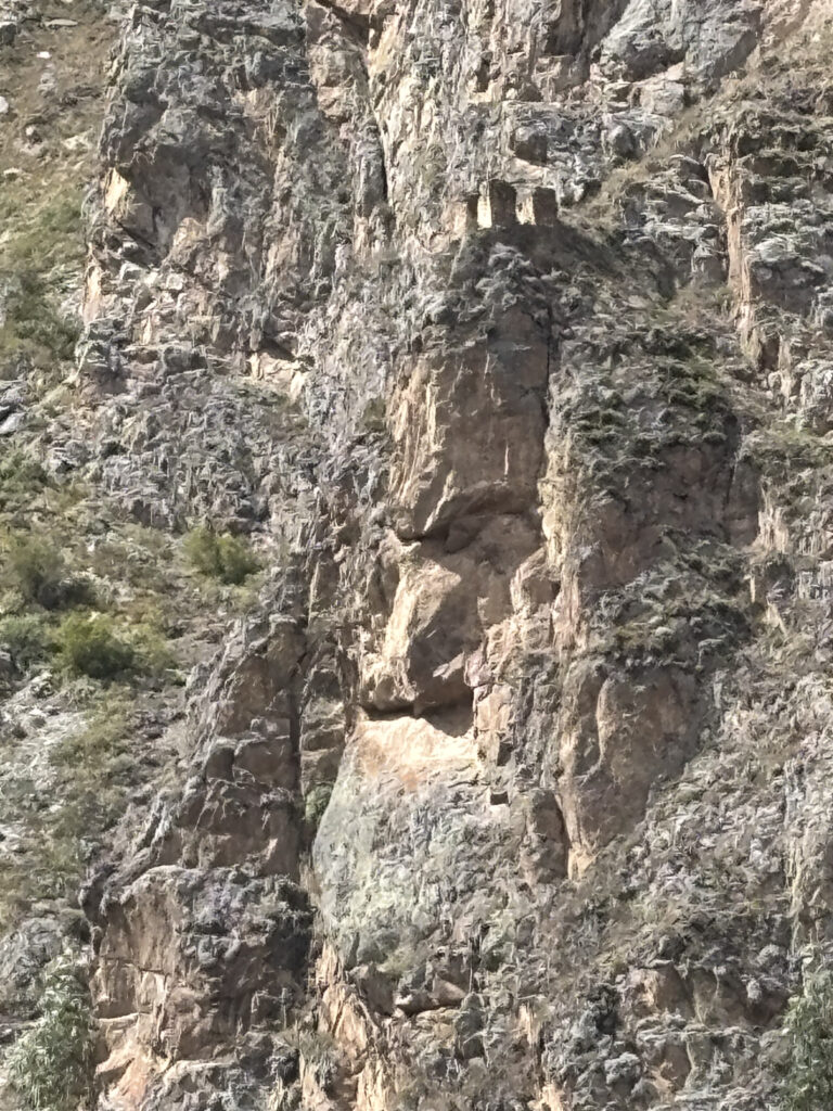 The image shows a rocky cliff face with natural formations. The surface is rugged and uneven, with patches of vegetation growing in some areas. The rocks have various shades of gray and brown, and there are some shadows cast on the cliff.