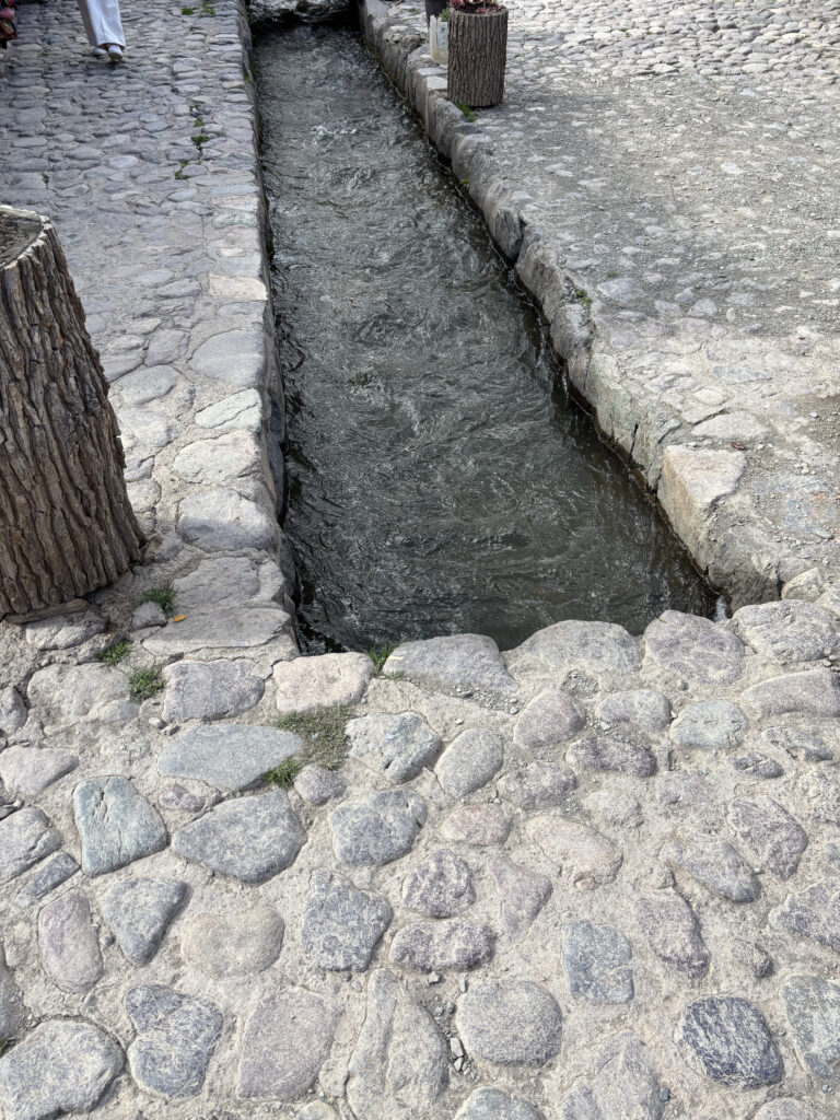 A narrow stone-lined canal with flowing water runs through a cobblestone pathway. The path is bordered by stone walls, and there are tree stump planters on either side. A person wearing white pants is partially visible walking on the left side of the image.