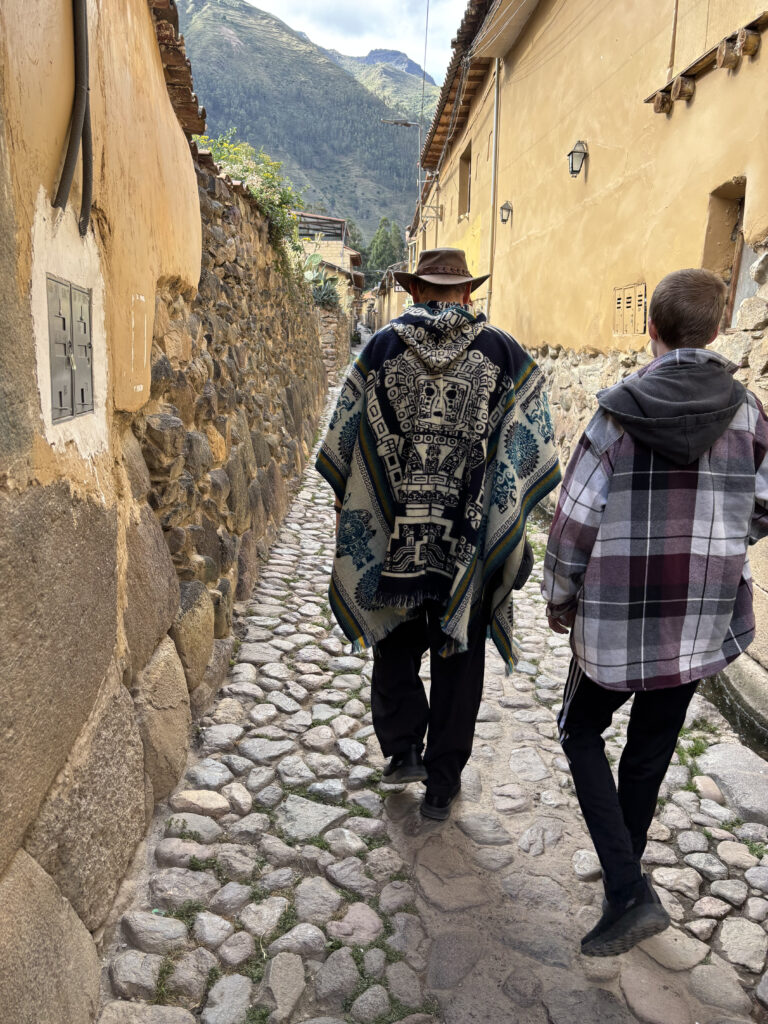 Two people are walking down a narrow cobblestone alley between stone and plaster walls. One person is wearing a patterned poncho and a wide-brimmed hat, while the other is in a plaid jacket. The alley is in a rustic setting with mountains visible in the background.