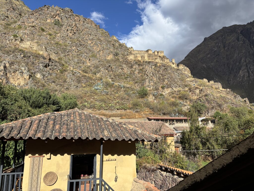 A rustic building with a tiled roof is in the foreground, featuring woven decorations on its wall. Behind it, a steep, rocky hillside is visible, with ancient stone ruins along the ridge. The sky is partly cloudy, with a mix of blue sky and dark clouds. Vegetation is sparse on the hillside, with some bushes and small plants.