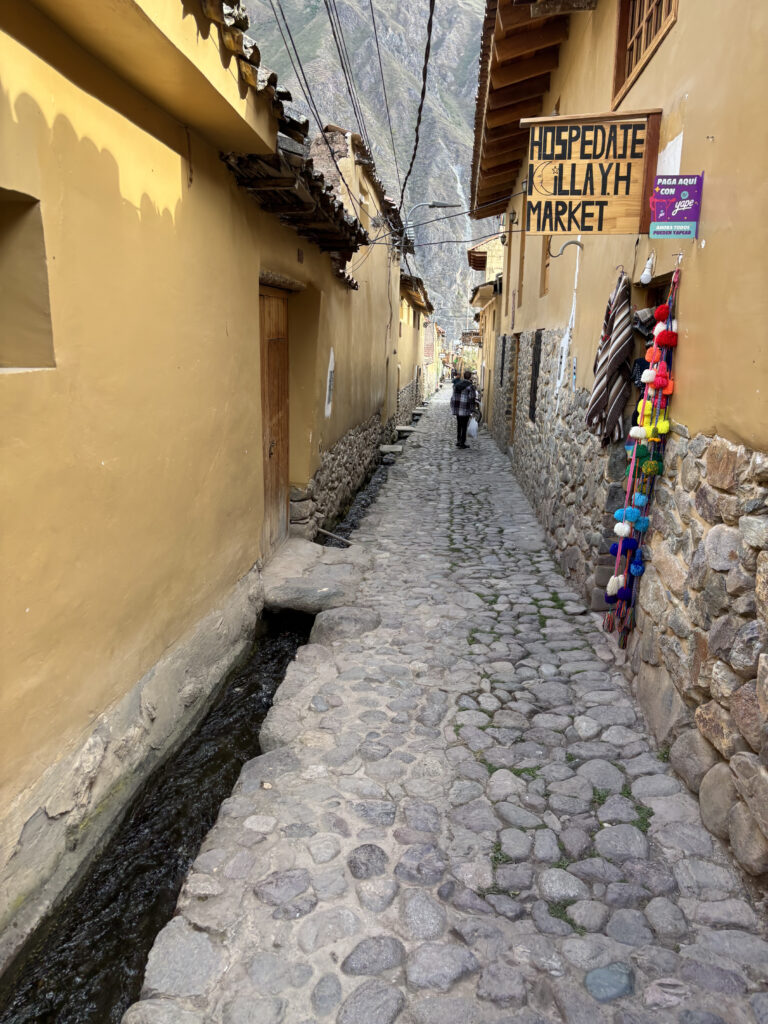 A narrow cobblestone alleyway with yellow and stone buildings on either side. A small water channel runs along the left side of the path. There is a sign on the right that reads "Hospedaje Killayh Market," and colorful textiles and pom-poms are displayed on the wall. Two people are walking in the distance. Overhead, wires are strung between the buildings, and mountains are visible in the background.