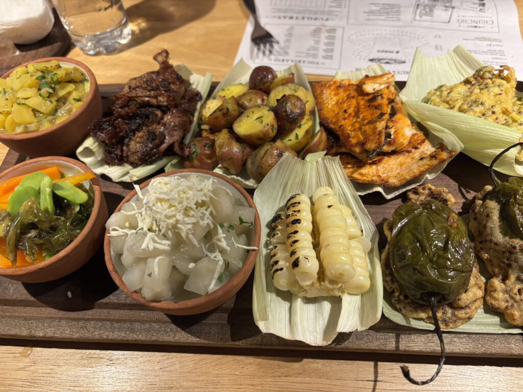 A wooden platter filled with a variety of foods, including grilled corn, roasted potatoes, grilled meat, and several small bowls containing different side dishes like pickled vegetables and diced potatoes. There are also stuffed peppers and a piece of grilled fish, all arranged on corn husks. A glass of water and a menu are visible in the background.
