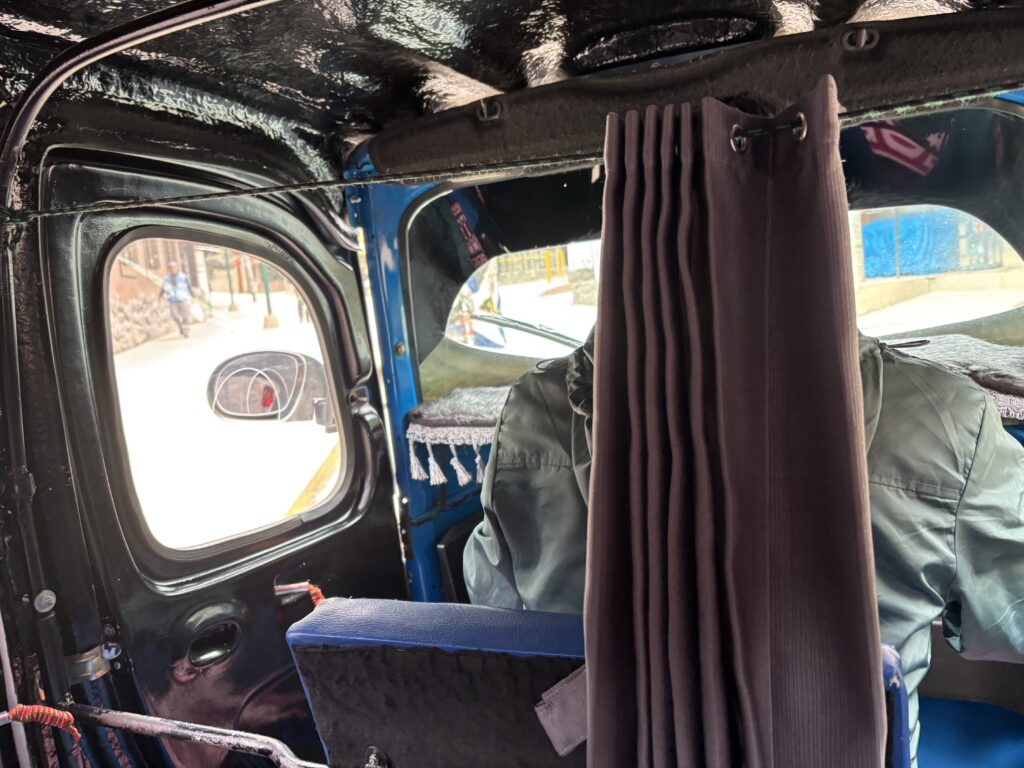 The image shows the interior of a small vehicle, possibly a rickshaw or tuk-tuk. There is a dark curtain hanging behind the driver's seat, partially obscuring the view. The vehicle's interior is black with some blue elements, and a side mirror is visible through the window. Outside, a street scene with a person walking on the sidewalk can be seen.