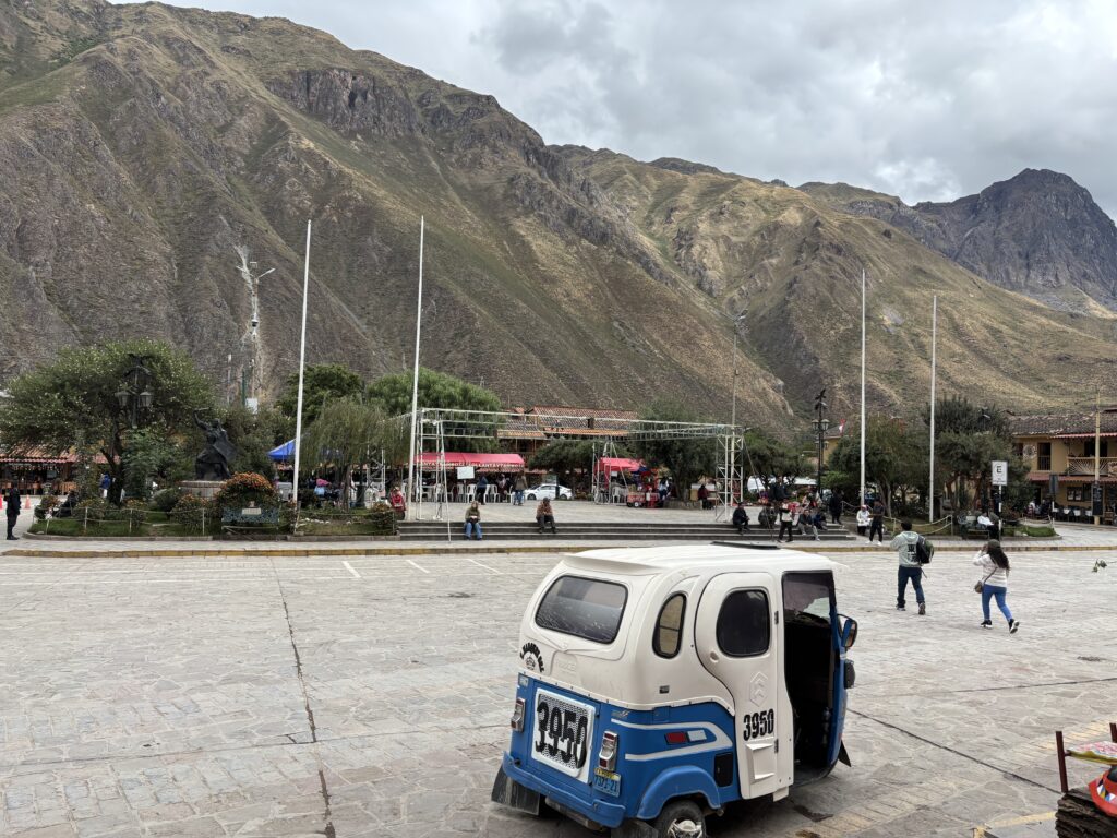 A small plaza is set against a backdrop of large, rugged mountains under a cloudy sky. In the foreground, a blue and white tuk-tuk is parked on a stone-paved area. The plaza features a statue surrounded by flowers and trees, with several flagpoles nearby. People are walking and sitting around the plaza, and there are market stalls with colorful canopies in the background.