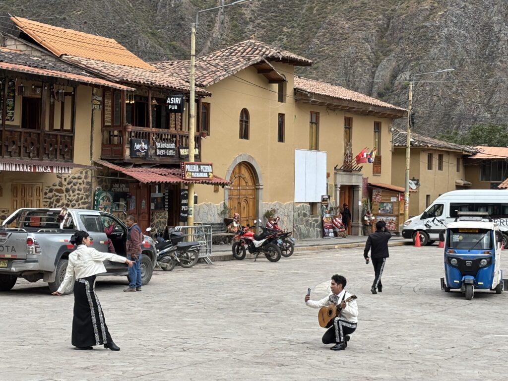 A street scene in a small town with traditional architecture. A person is kneeling on the ground playing a guitar, while another person stands nearby, posing with an arm outstretched. There are several vehicles, including a pickup truck, motorcycles, a van, and a tuk-tuk. The buildings have wooden balconies and signs for a pub and a restaurant. The background features rocky hills.