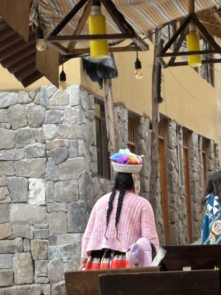 A person with long braided hair is wearing a colorful traditional outfit, including a pink sweater and a decorative hat with flowers. They are standing near a stone building with wooden windows. Above them, there are hanging light bulbs and a striped canopy. Another person is partially visible on the right, wearing a patterned garment.