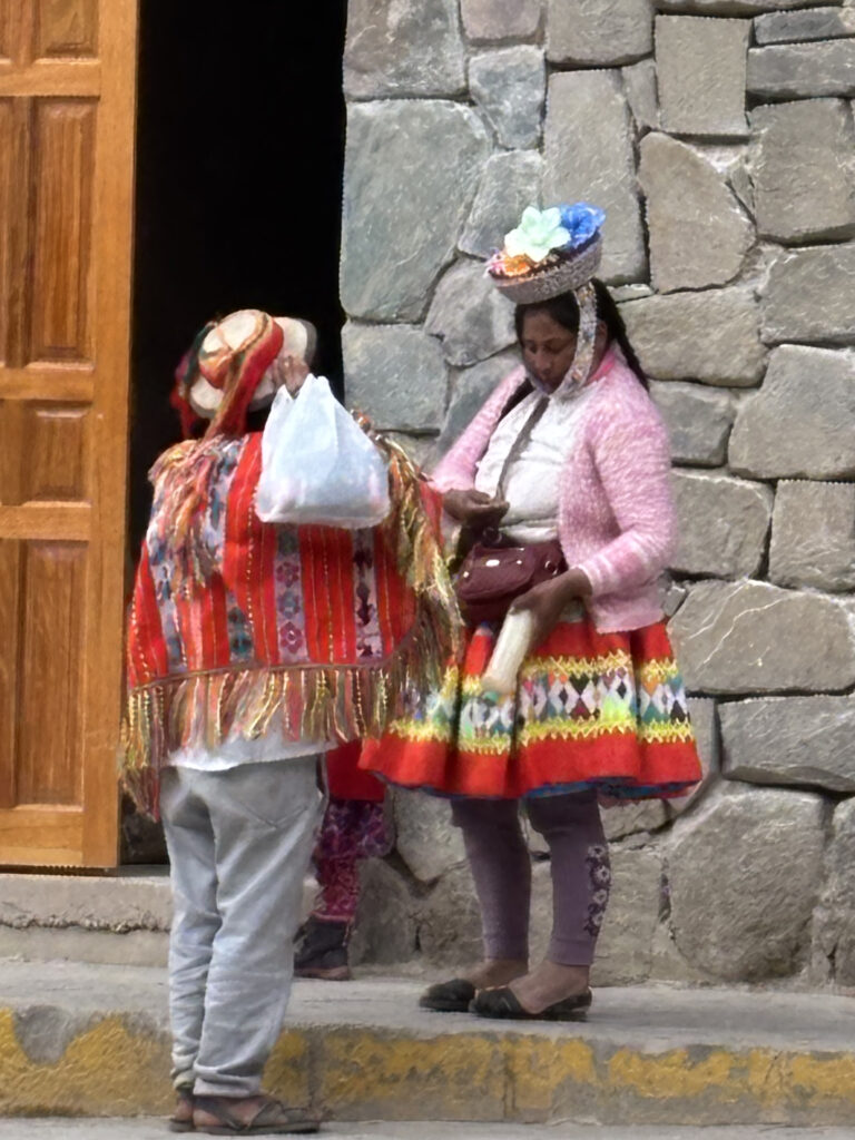 Two people are standing in front of a stone wall. They are wearing traditional, colorful clothing with intricate patterns. One person is wearing a hat adorned with flowers and is holding a bag. The other person has a white bag over their shoulder. The scene appears to be in a cultural or traditional setting.