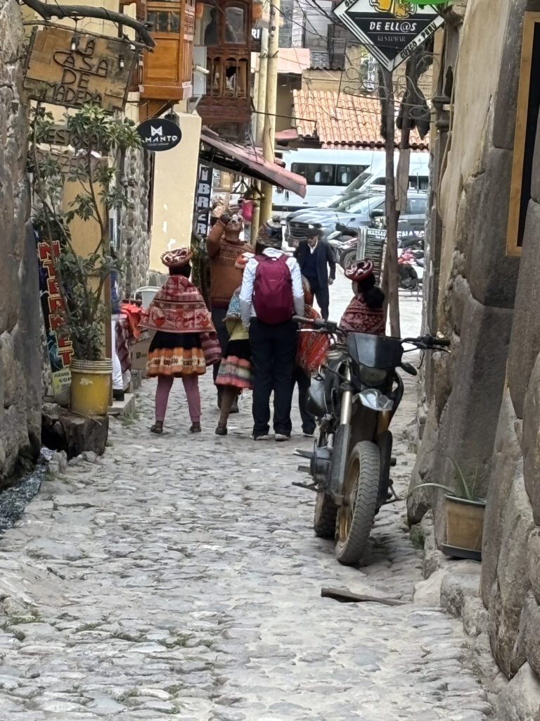A narrow cobblestone street with a group of people wearing traditional colorful clothing. They are walking away from the camera. A motorcycle is parked on the right side of the street. The buildings are made of stone and have signs and plants along the walls. The street is lined with rustic architecture, and there are various signs in Spanish.