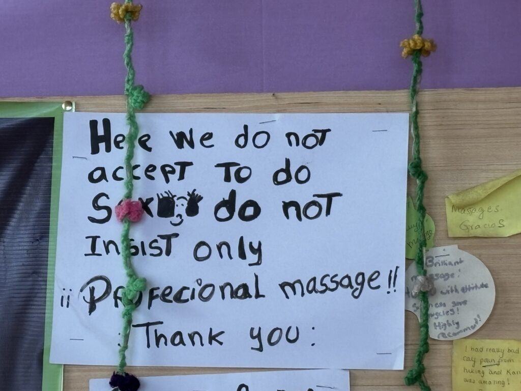The image shows a handwritten sign pinned to a board. The sign reads: "Here we do not accept to do sex, do not insist, only professional massage!! Thank you." The sign is decorated with a green yarn string with small flowers attached. There are also small notes pinned to the board around the sign.