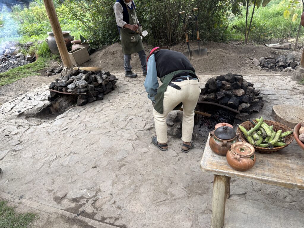 The image shows two people working outdoors near a traditional cooking setup. There are two stone-lined pits with fires, and one person is tending to one of the pits. A wooden table in the foreground holds clay pots and a basket of green vegetables. In the background, there are shovels, a broom, and some large clay pots. The area is surrounded by greenery.