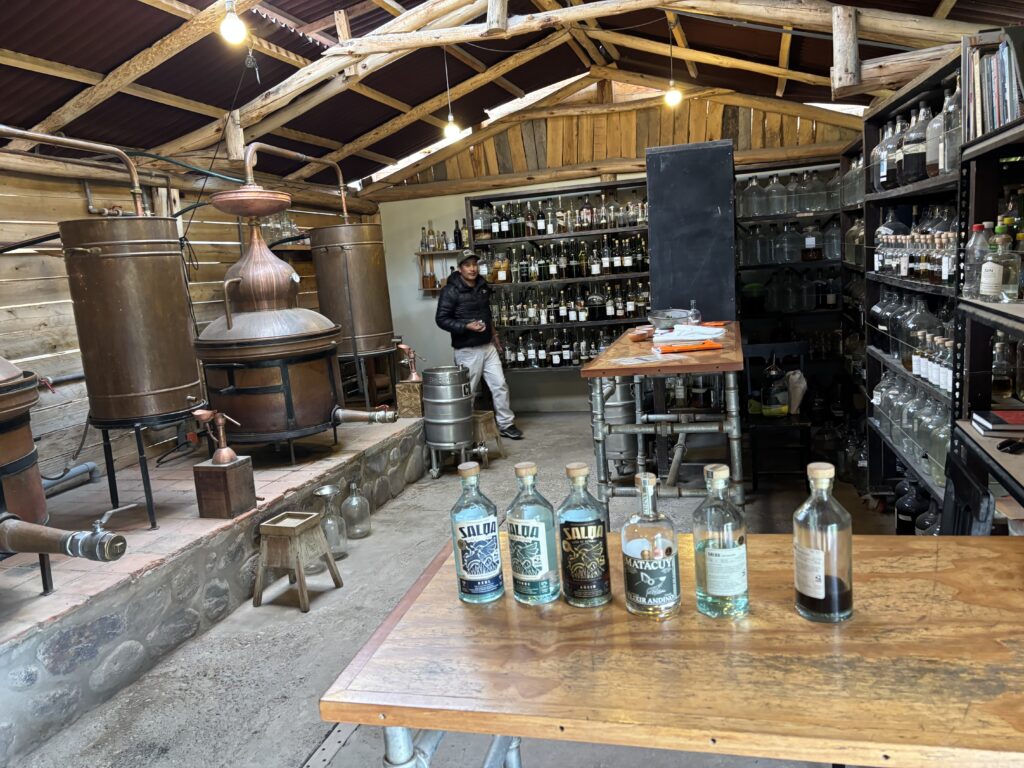 The image shows the interior of a distillery. There are several large copper stills on the left side, used for distillation. In the foreground, a wooden table displays several bottles of spirits. Shelves filled with various bottles line the walls in the background. A person is standing near the shelves, and the room has a rustic wooden ceiling with exposed beams.