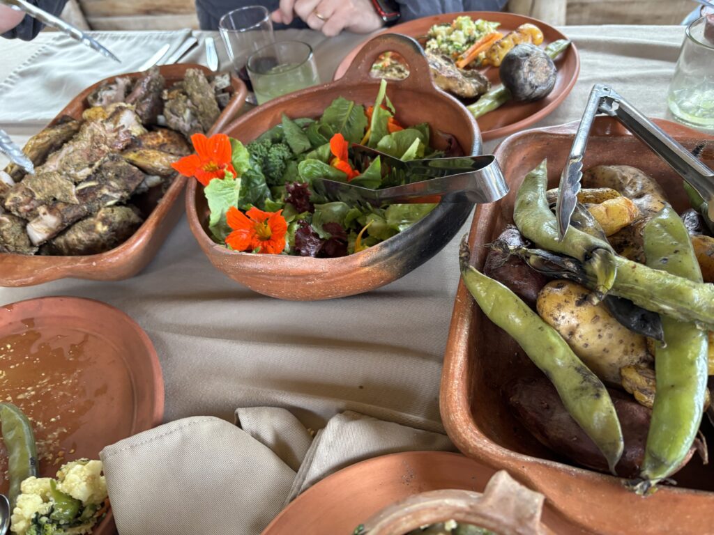 A table is set with various dishes in clay pots. One pot contains grilled or roasted meat, another has a fresh salad with leafy greens and bright orange edible flowers. A third pot holds a mix of roasted potatoes and green beans. There are also plates with additional food items, a glass with a drink, and utensils on the table. The setting appears to be rustic and inviting.