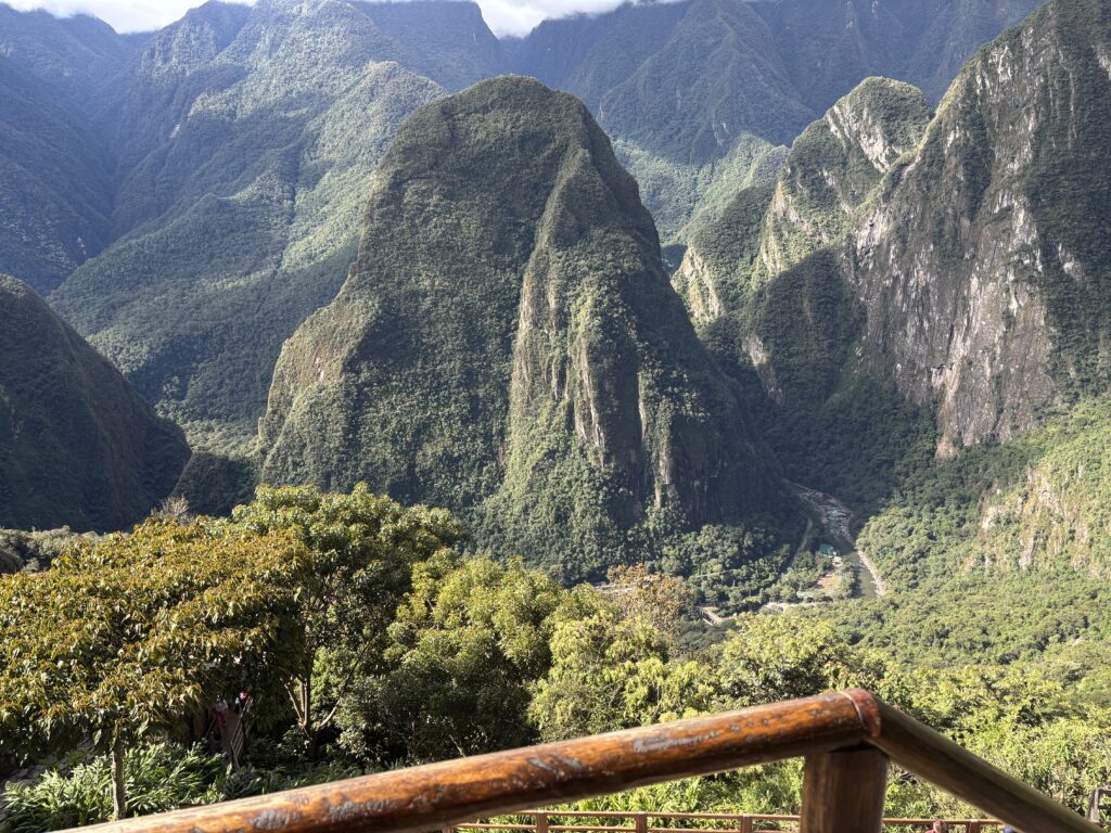 A scenic view of lush, green mountains with steep cliffs and dense vegetation. In the foreground, there is a wooden railing, and below, a river winds through the valley. The sky is partly cloudy, casting shadows on the landscape.
