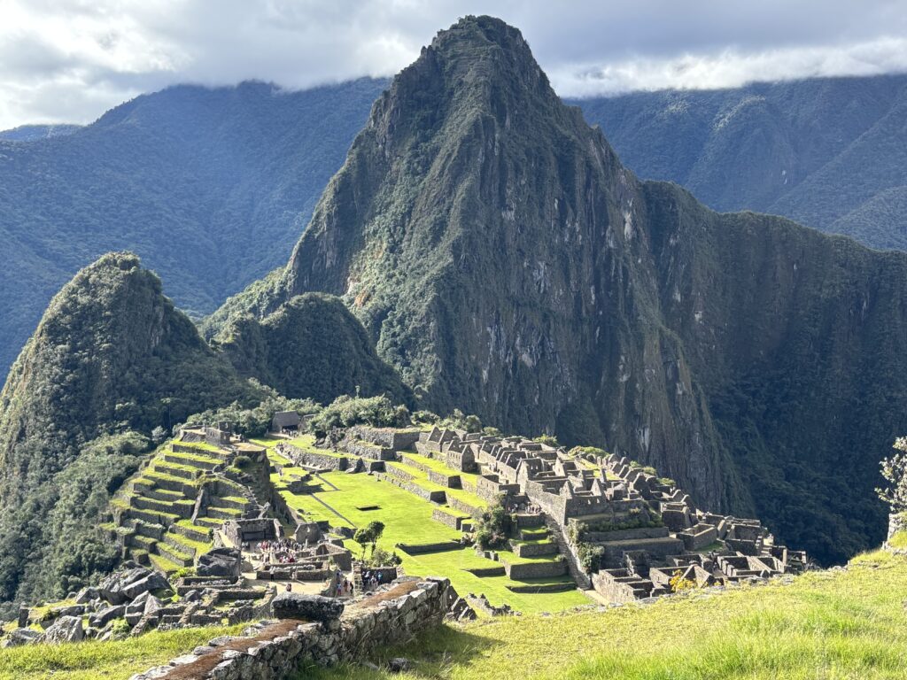 The image shows the ancient Incan city of Machu Picchu, located in the Andes Mountains of Peru. The site features terraced fields and stone structures set against a backdrop of steep, lush green mountains. The sky is partly cloudy, casting shadows over the landscape.