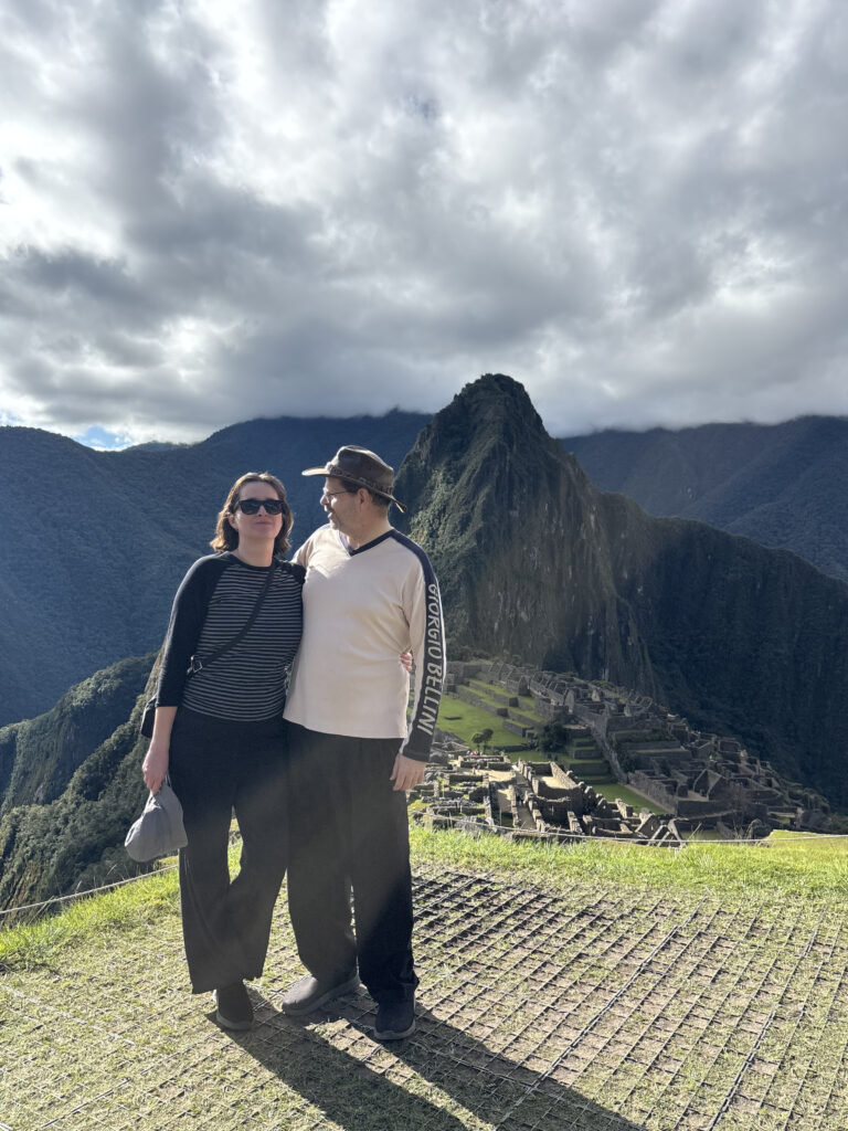 A couple is standing on a grassy area with Machu Picchu in the background. The sky is cloudy, and the mountains are visible behind the ancient ruins. The person on the left is wearing sunglasses and a striped shirt, while the person on the right is wearing a hat and a light-colored shirt.