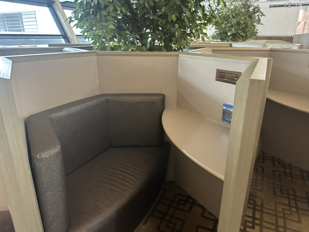 The image shows a small, semi-enclosed seating area with a cushioned bench and a curved desk surface. There is a power outlet on the wall above the desk. The area is surrounded by light-colored partitions, and there is a large potted plant nearby. The setting appears to be in a public or office space with a patterned carpet on the floor.