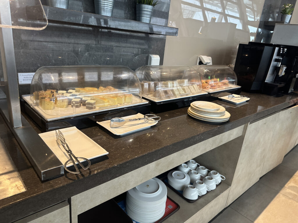 The image shows a buffet counter with a variety of food items displayed under clear plastic covers. There are different types of pastries and sandwiches. In front of the food, there are serving utensils on small plates. To the right, there is a stack of plates and a coffee machine. Below the counter, there are shelves with stacked cups and bowls. The background features a dark wall with potted plants on a shelf.