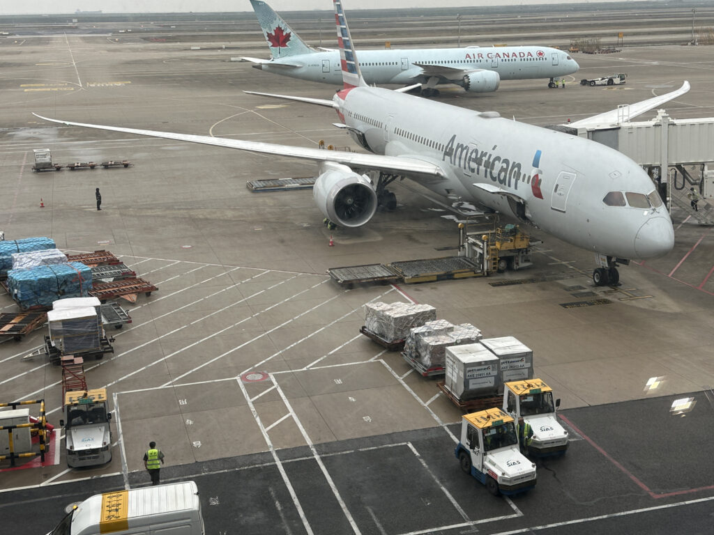 The image shows an airport tarmac with two airplanes. In the foreground, an American Airlines plane is parked at a gate, with cargo and ground support vehicles nearby. In the background, an Air Canada plane is visible on the tarmac. The scene includes various airport personnel and equipment, such as luggage carts and cargo pallets. The sky appears overcast.