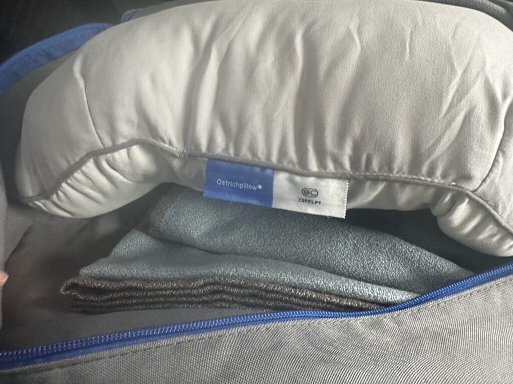 The image shows a close-up of a travel pillow with a label that reads "Ostrichpillow." The pillow is light gray and appears to be inside a bag with a blue zipper. There is also a folded fabric or blanket beneath the pillow.