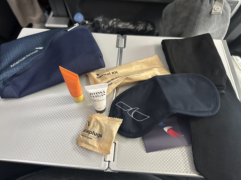 The image shows a collection of travel amenities on an airplane tray table. Items include a navy blue zippered pouch with an airline logo, a small tube of orange cream, a tube of hand cream, a packet labeled "Dental Kit," a pair of earplugs in a paper wrapper, a navy blue eye mask, and a pair of black socks. There is also a card with a logo partially visible.