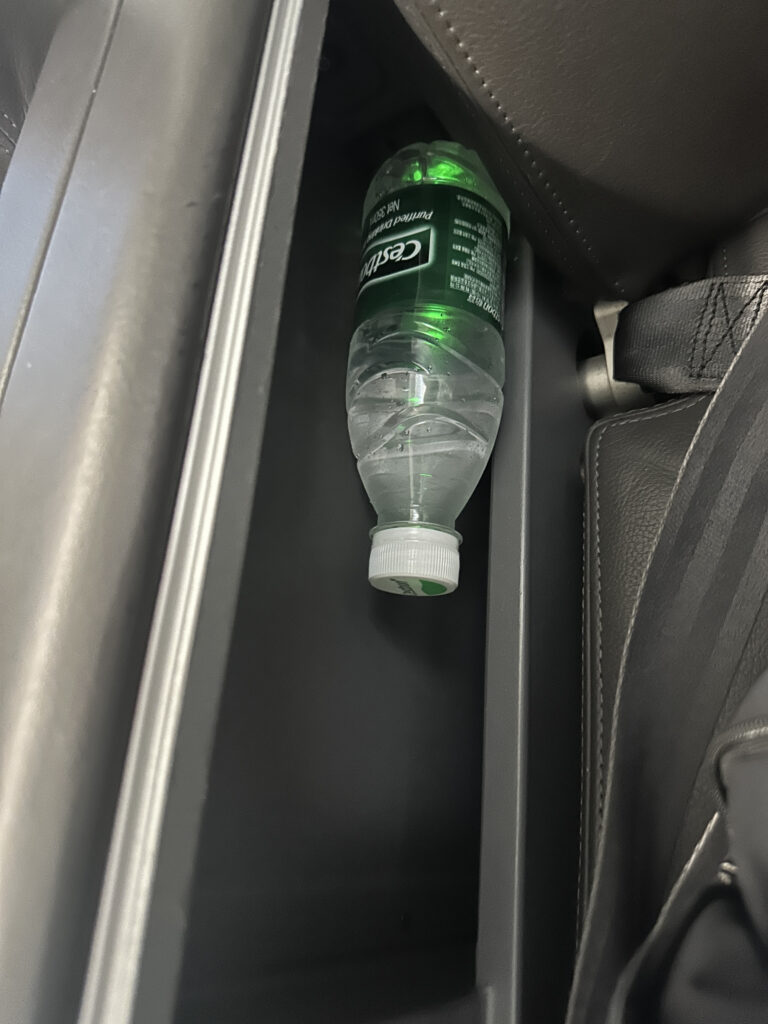 A plastic water bottle is wedged between the seat and the center console of a car. The bottle is positioned upside down, with the cap facing upwards. The interior of the car is visible, showing parts of the seat and console.