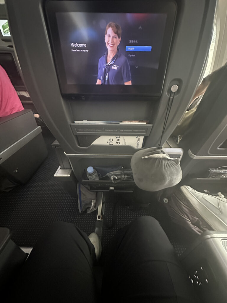 The image shows the interior of an airplane from a passenger's perspective. A screen on the back of the seat in front displays a welcome message with a flight attendant and language selection options. Below the screen, there is a pocket containing a water bottle and other items. The passenger's legs and feet are visible, and there is a gray neck pillow hanging on the seat in front. The cabin appears to be in a business or first-class section.