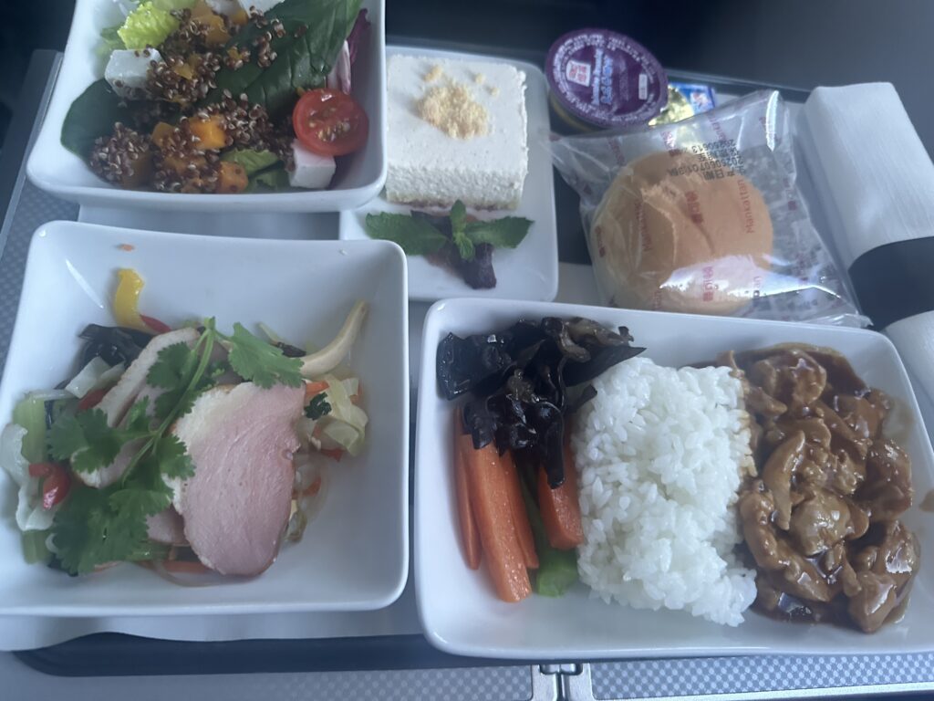 The image shows an airplane meal consisting of several dishes. There is a salad with mixed greens, cherry tomatoes, and quinoa. Another dish contains slices of meat with vegetables and cilantro. A main dish features rice, glazed carrots, and a meat dish with sauce. There is also a dessert that appears to be a slice of cake, a packaged bread roll, and a small container of butter. A napkin and utensils are placed on the side.