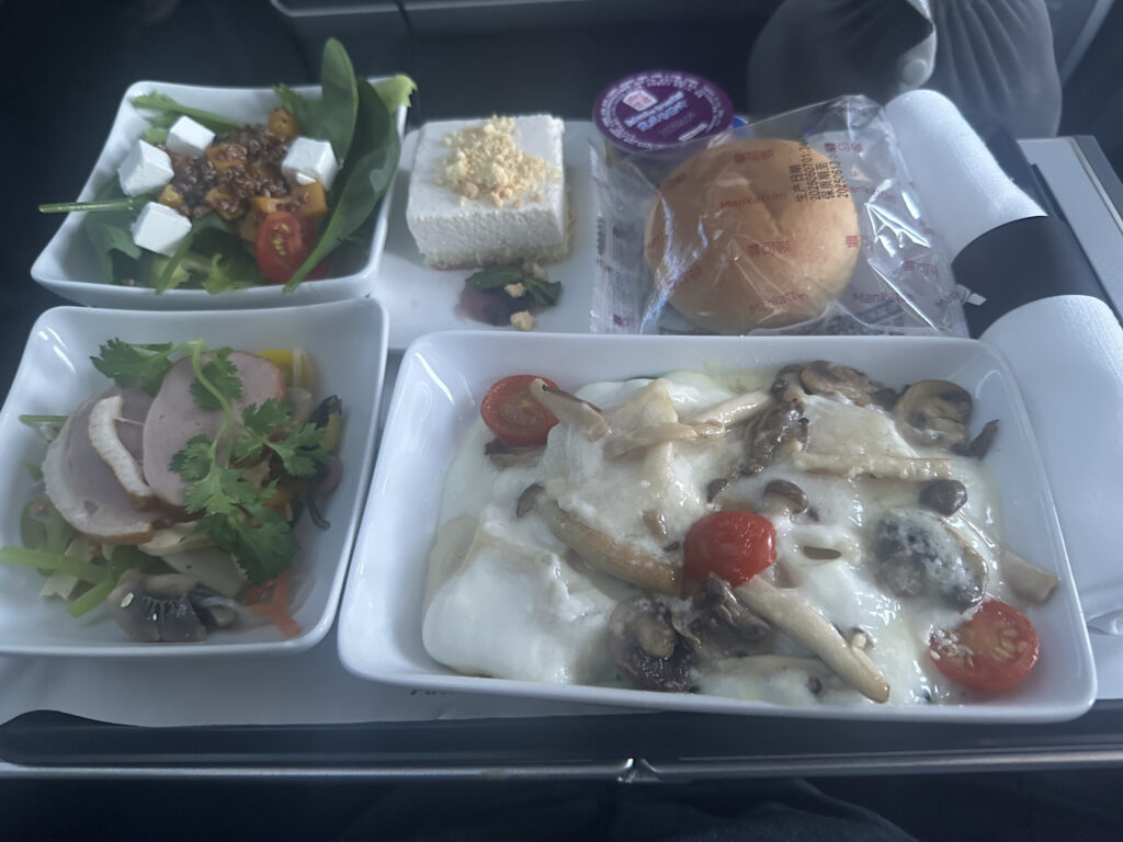 The image shows an airplane meal consisting of several dishes. In the foreground, there is a main dish with melted cheese, mushrooms, and cherry tomatoes. To the left, there is a small bowl with slices of meat garnished with cilantro and vegetables. Above that, there is a salad with spinach, cherry tomatoes, feta cheese cubes, and quinoa. Next to the salad, there is a dessert square topped with crumbled nuts. In the background, there is a packaged bread roll and a small container of butter or spread, along with a rolled napkin and cutlery.