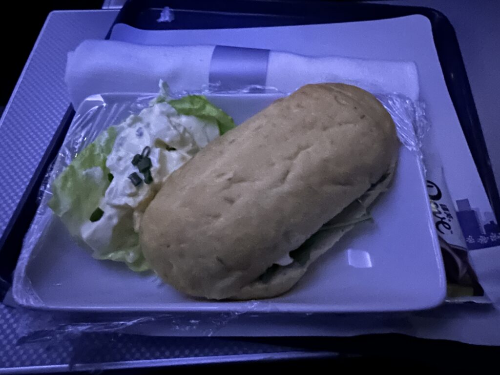 A meal on a tray featuring a sandwich and a side of potato salad with chives on a lettuce leaf. The tray also includes a napkin and a packaged item, possibly a snack or dessert.