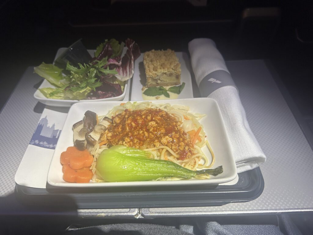 The image shows an airline meal on a tray. The main dish consists of noodles topped with a spicy sauce, mushrooms, carrot slices, and a piece of bok choy. There is a side salad with mixed greens and a dessert that appears to be a layered cake. A rolled-up napkin with a utensil is placed next to the dishes.